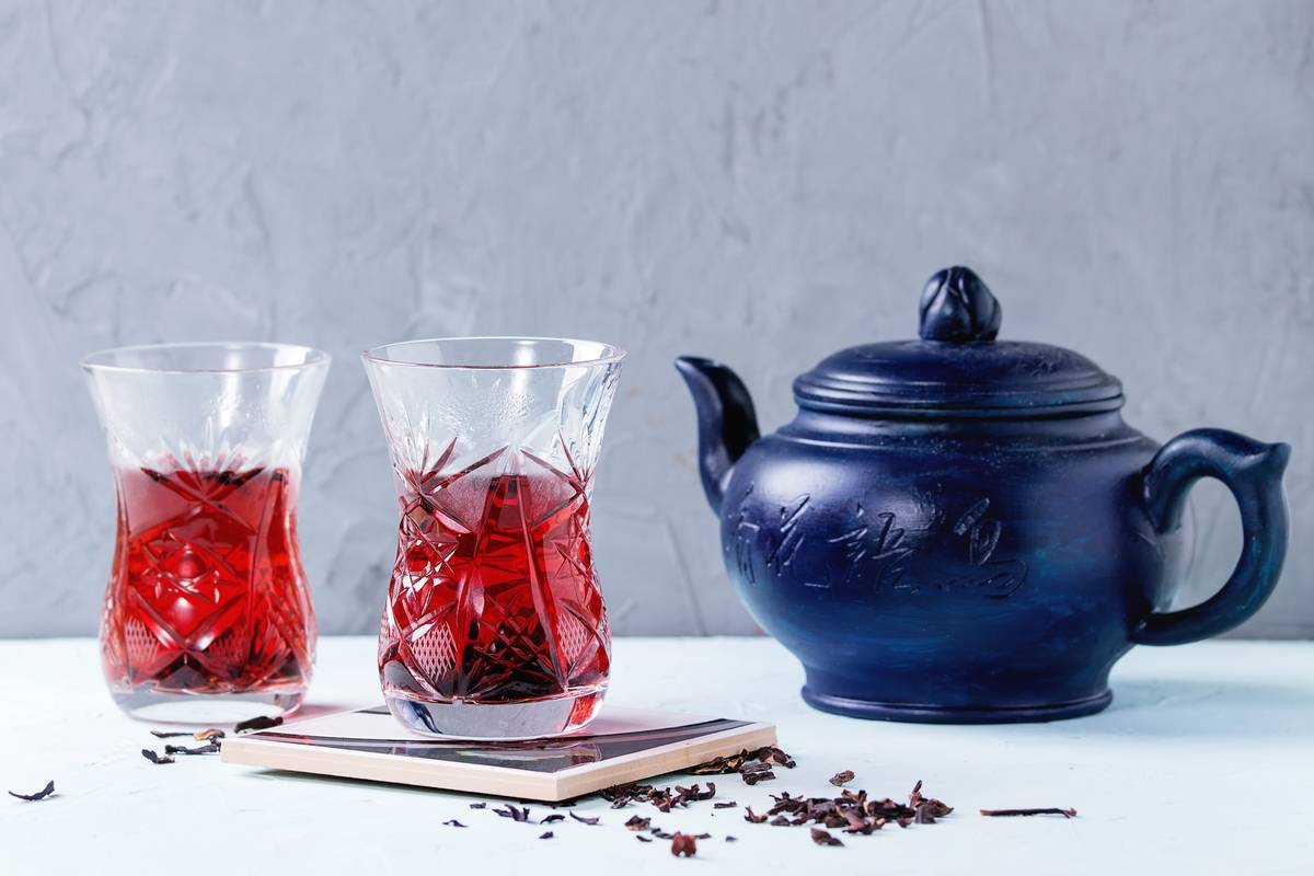 Two traditional asian style glasses with hibiscus tea karkade, served with dry hibiskus and dark blue ceramic teapot over blue and gray textured background