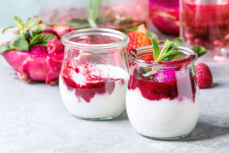 Jars of natural white yogurt with berry sauce, fruit salad with pink  dragon fruit, berries and mint, served with bottle of lemonade on grey 
table Close up Healthy eating.
