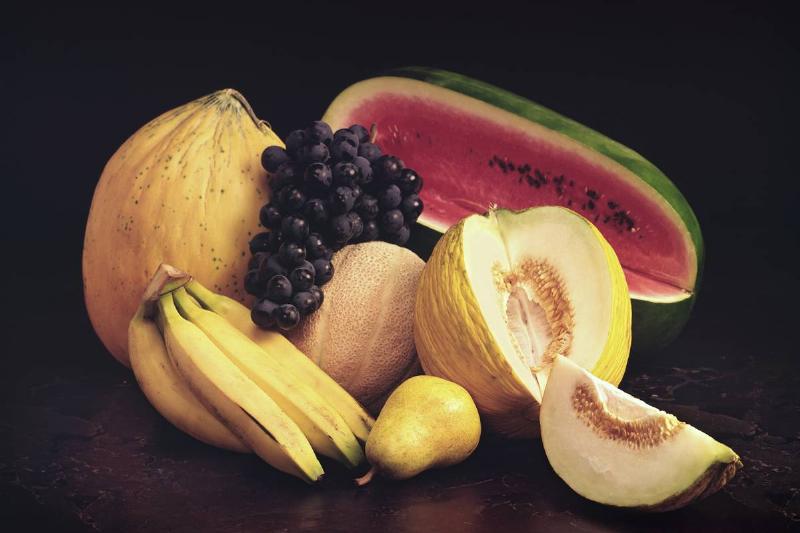 View of a several of melons (watermelon, cantalope, honeydew, Galia),  grapes, bananas, and a pear of a black marble table top, 1990.