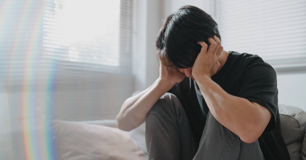 A man sat on his bed, knees drawn to his chest, clutching his head with his hands.