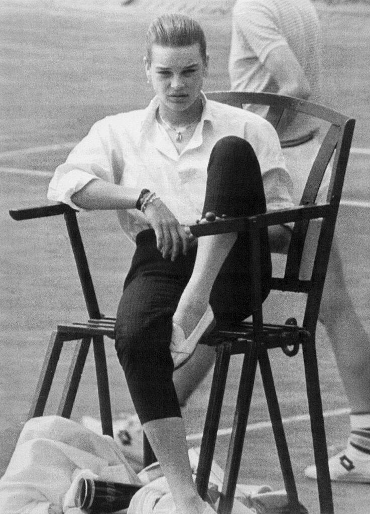 Princess Stéphanie sitting in a chair, while a man walks by behind her